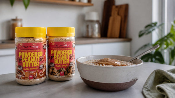 Two Macro Mike powdered peanut butter jars beside a bowl of porridge drizzled with peanut sauce on a kitchen bench