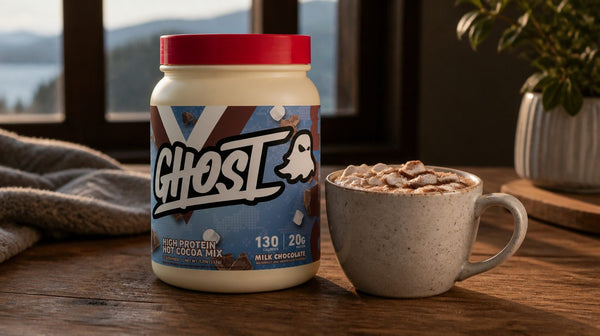 Mug of hot chocolate topped with mini marshmallows beside a bag of Ghost High Protein Hot Cocoa on a wooden table