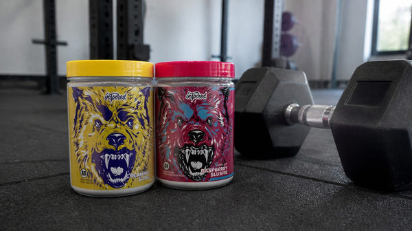Inspired DVST8 BBD Creaming Soda and Raspberry Slushie pre-workout tubs on a black gym floor beside a dumbbell