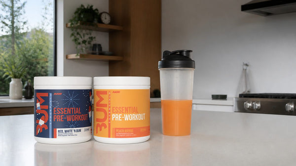 CBUM Essential White N Burn and Peach Mango pre-workout tubs beside a shaker of orange drink