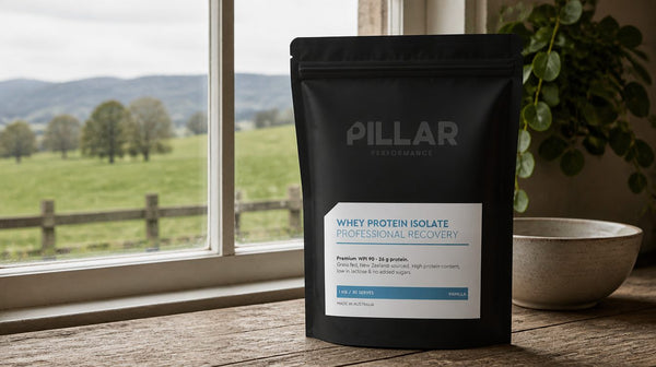 Pillar Whey Protein Isolate tub on a farmhouse bench with green New Zealand pasture visible through the window