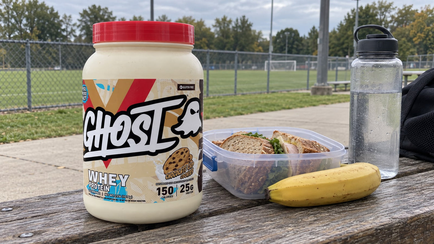 GHOST Whey Chocolate Chip Cookies beside a packed lunch and water at a sports field.