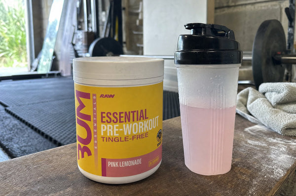A tub of RAW Essential Pre-Workout Tingle-Free in Pink Lemonade on a wooden bench in a home garage gym in early morning light